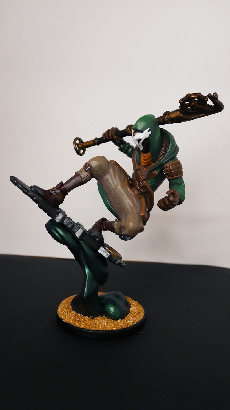 Pu&ograve; includere: Statuetta di un personaggio in posa dinamica, che indossa un abito con cappuccio verde con pantaloni e stivali marroni. La figura tiene in mano un bastone ed &egrave; montata su una base verde scuro con una superficie strutturata color oro.