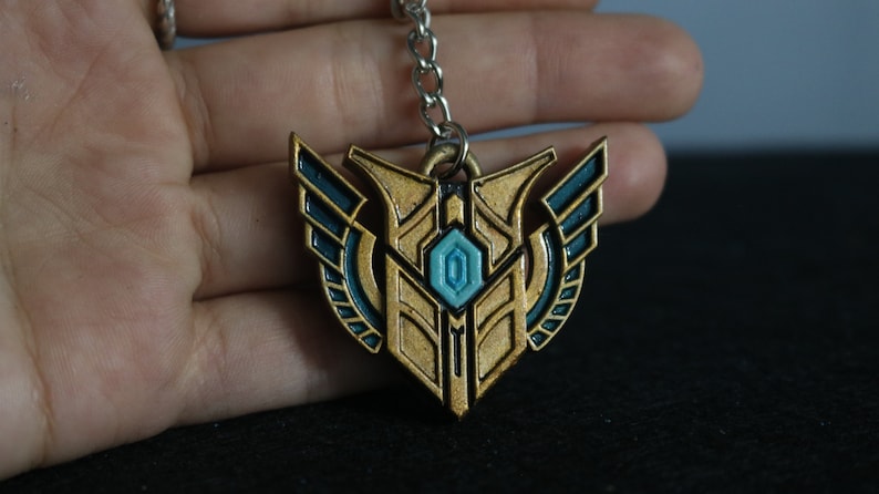 Op de afbeelding: Een gouden en blauwgroene metalen sleutelhangerhanger. De hanger heeft de vorm van een schild met een centrale blauwgroene edelsteen en blauwgroene accenten. Hij hangt aan een zilveren ketting.