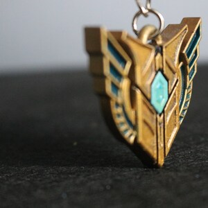 Op de afbeelding: Een goudkleurige hanger met turquoise accenten en een centrale turquoise edelsteen. De schildvormige hanger heeft een gedetailleerd ontwerp en is bevestigd aan een zilverkleurige ketting. Het is waarschijnlijk een sieraad of een decoratief accessoire.