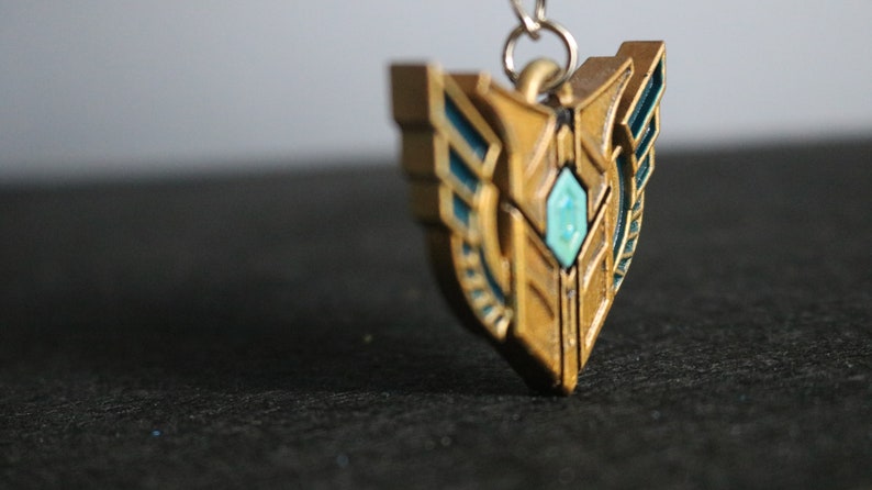Op de afbeelding: Een goudkleurige hanger met turquoise accenten en een centrale turquoise edelsteen. De schildvormige hanger heeft een gedetailleerd ontwerp en is bevestigd aan een zilverkleurige ketting. Het is waarschijnlijk een sieraad of een decoratief accessoire.