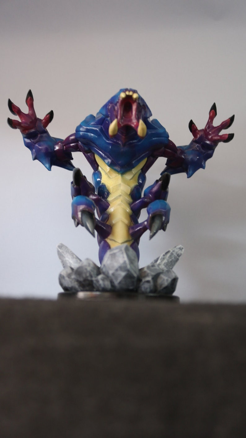 May include: A detailed figurine of a fantasy creature with a blue and purple armored body, yellow underbelly, and red mouth. The figure has outstretched arms with clawed hands and is mounted on a rocky base.