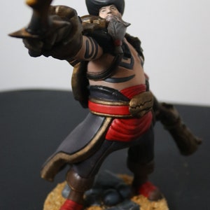 May include: A detailed figurine of a pirate character, posed with a curved blade. The figure wears a black hat, a red sash, and brown boots. The base is textured with sand and small rocks. The pirate has a beard and tattoos.