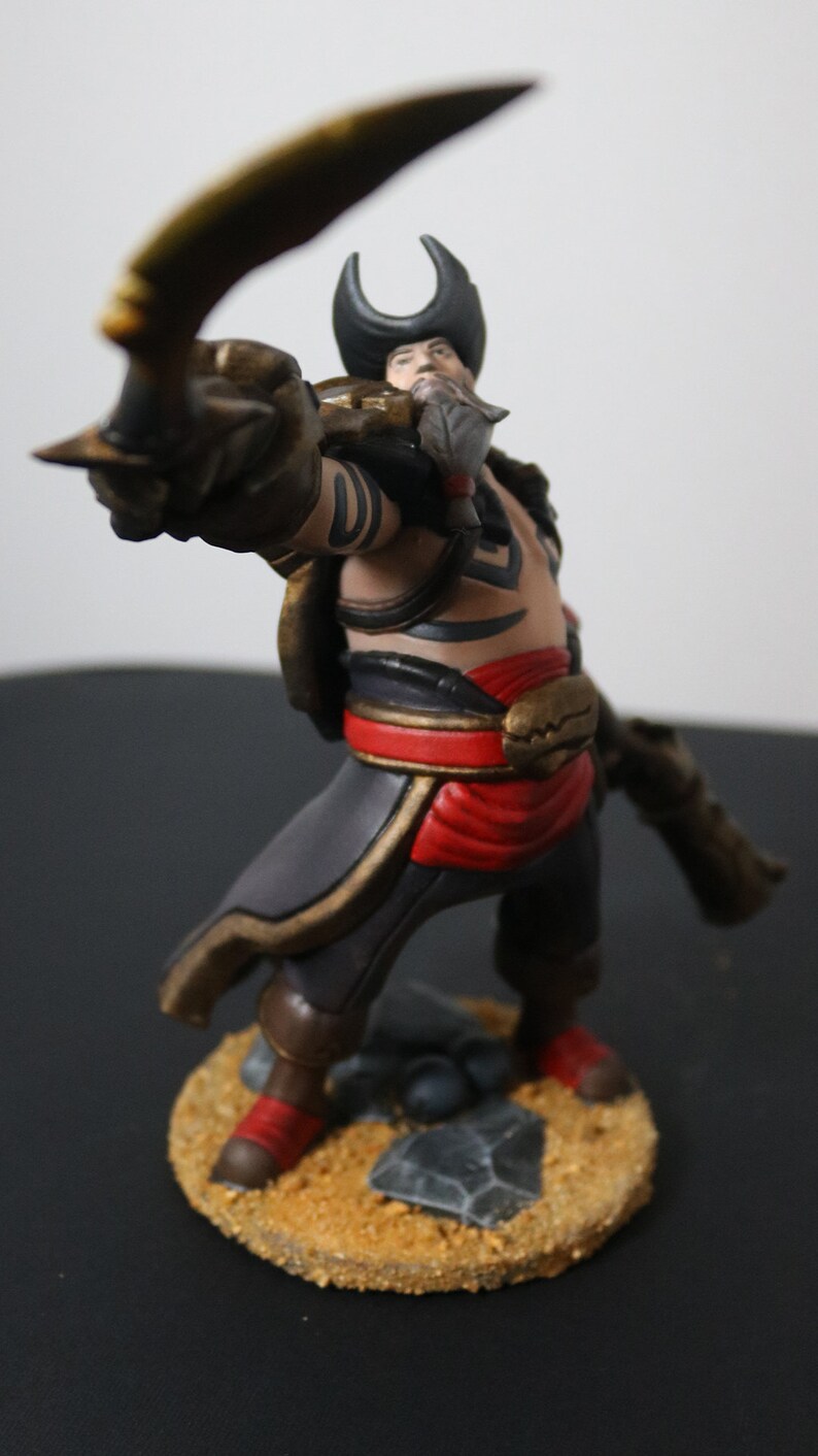 May include: A detailed figurine of a pirate character, posed with a curved blade. The figure wears a black hat, a red sash, and brown boots. The base is textured with sand and small rocks. The pirate has a beard and tattoos.
