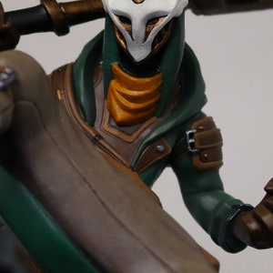 Pu&ograve; includere: Statuetta dettagliata di un personaggio con un mantello con cappuccio verde e una maschera bianca. La figura ha accenti dorati sulla maschera e sull'armatura, con un abito beige e marrone. La figura tiene un bastone.