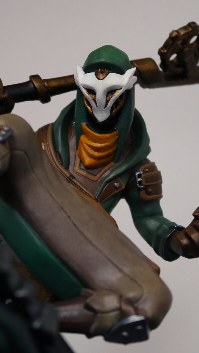 Pu&ograve; includere: Statuetta dettagliata di un personaggio con un mantello con cappuccio verde e una maschera bianca. La figura ha accenti dorati sulla maschera e sull'armatura, con un abito beige e marrone. La figura tiene un bastone.