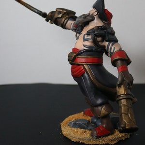 May include: A detailed figurine of a pirate with a sword raised. The figure wears a black hat, red sash, and dark pants. The base is textured with sand and small rocks. The pirate has tattoos and a beard.
