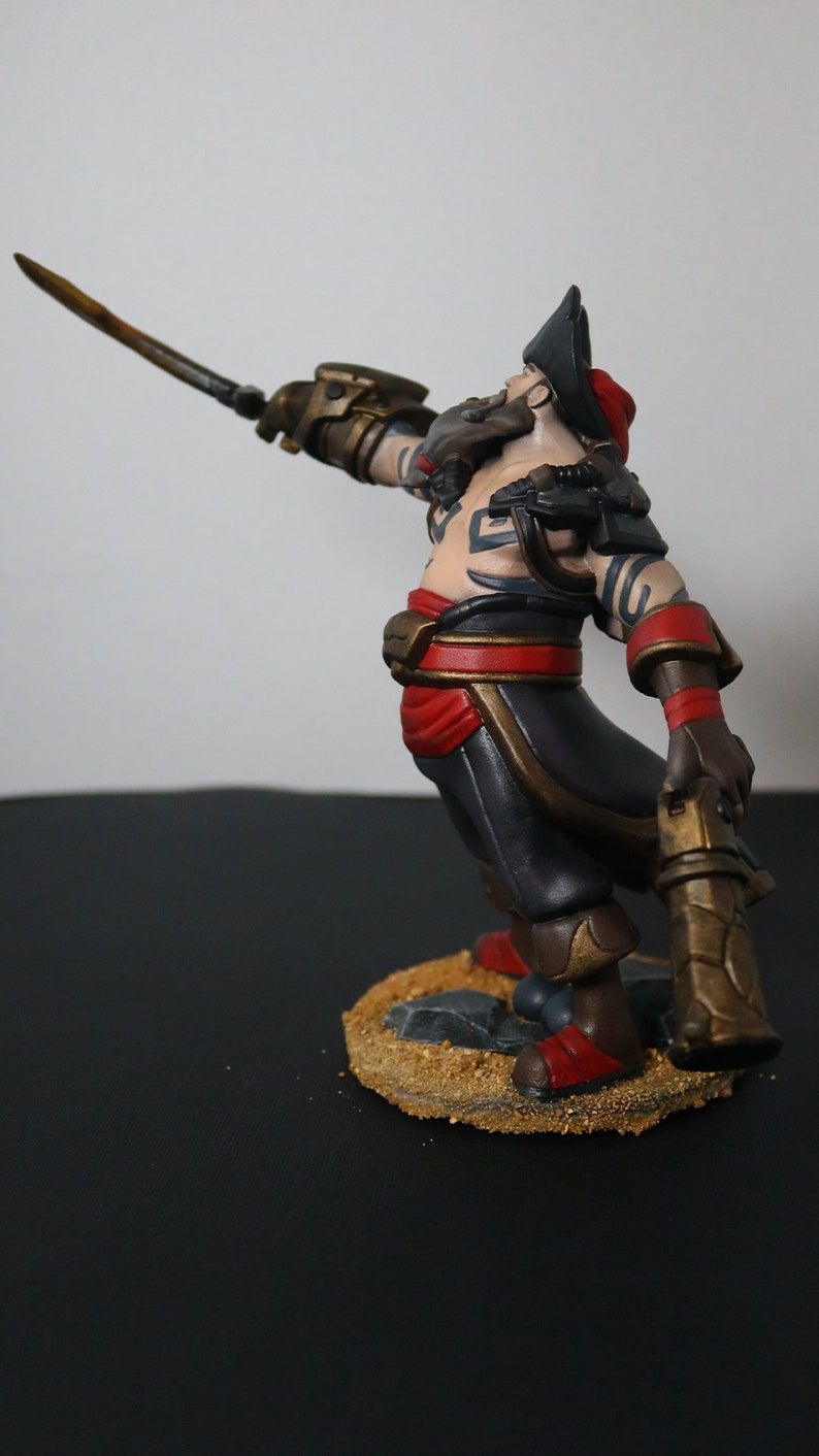 May include: A detailed figurine of a pirate with a sword raised. The figure wears a black hat, red sash, and dark pants. The base is textured with sand and small rocks. The pirate has tattoos and a beard.