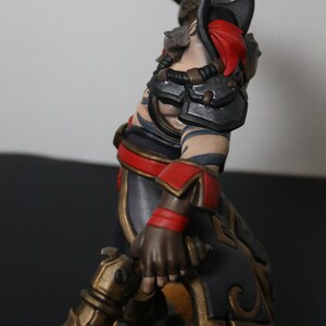 May include: A detailed figurine of a warrior in a dynamic pose, wielding a sword and a decorative weapon. The figure features a color scheme of black, red, and gold accents, with intricate armor and tattoos. The base is textured with sand-like material.