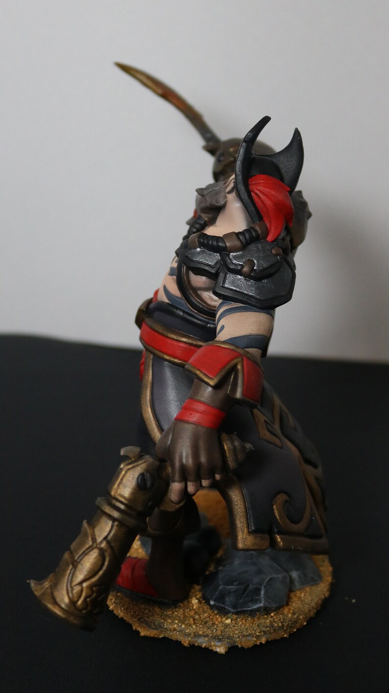 May include: A detailed figurine of a warrior in a dynamic pose, wielding a sword and a decorative weapon. The figure features a color scheme of black, red, and gold accents, with intricate armor and tattoos. The base is textured with sand-like material.