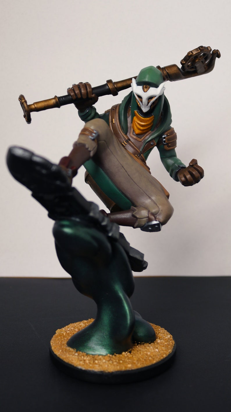 Pu&ograve; includere: Statuetta dettagliata di un personaggio in posa dinamica, con un bastone. La figura &egrave; vestita con abiti verdi, marroni e beige, con una maschera bianca. La base &egrave; scura con un aspetto strutturato e sabbioso.