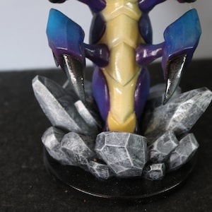 May include: A detailed figurine of a creature with a yellow and purple body, blue and purple claws, and a rocky base. The figurine is set on a black circular base, with the rocks in shades of gray.