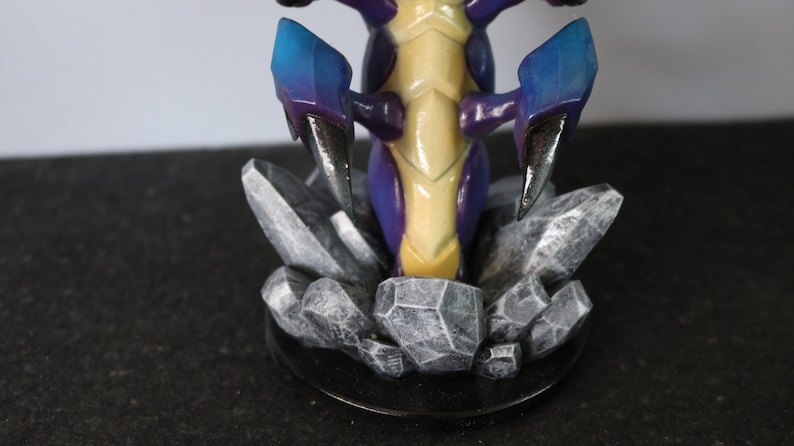 May include: A detailed figurine of a creature with a yellow and purple body, blue and purple claws, and a rocky base. The figurine is set on a black circular base, with the rocks in shades of gray.
