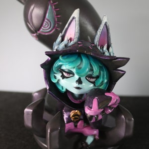 May include: A detailed figurine of a character with teal hair, wearing a dark hooded cloak with cat-like ears. The figure has a pale complexion and is holding a smaller, matching figure. A larger, shadowy figure looms behind.
