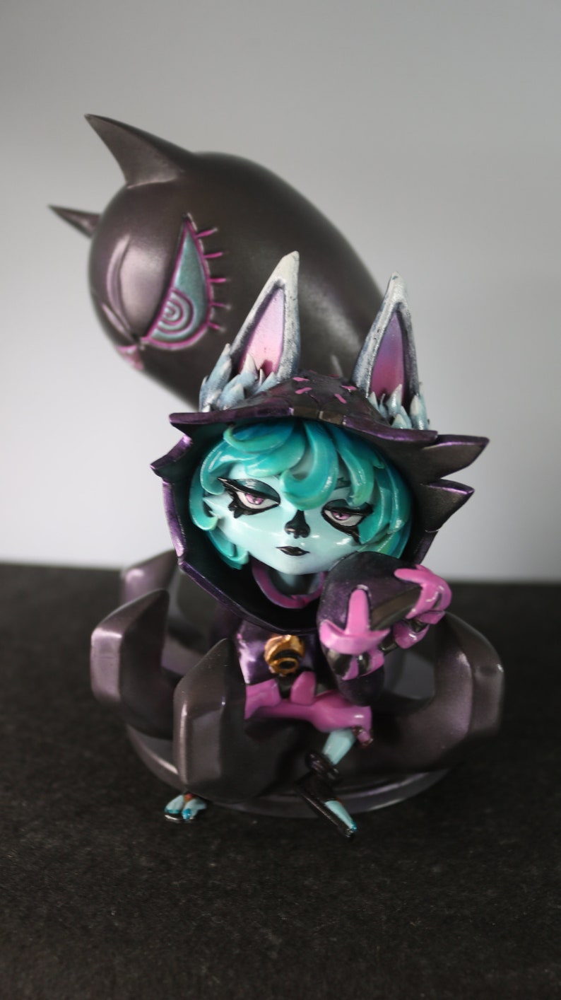 May include: A detailed figurine of a character with teal hair, wearing a dark hooded cloak with cat-like ears. The figure has a pale complexion and is holding a smaller, matching figure. A larger, shadowy figure looms behind.