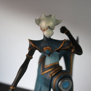 May include: A figurine of a stylized humanoid figure with a white head and a blue and black body. The figure has gold accents and is posed with a clenched fist. The figurine is likely a collectible or decorative item.