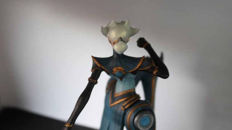 May include: A figurine of a stylized humanoid figure with a white head and a blue and black body. The figure has gold accents and is posed with a clenched fist. The figurine is likely a collectible or decorative item.