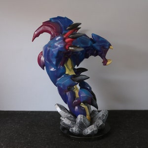 May include: A detailed figurine of a fantasy creature, predominantly blue with accents of red, yellow, and black. The sculpture is posed in an aggressive stance, mounted on a base of gray rock formations.