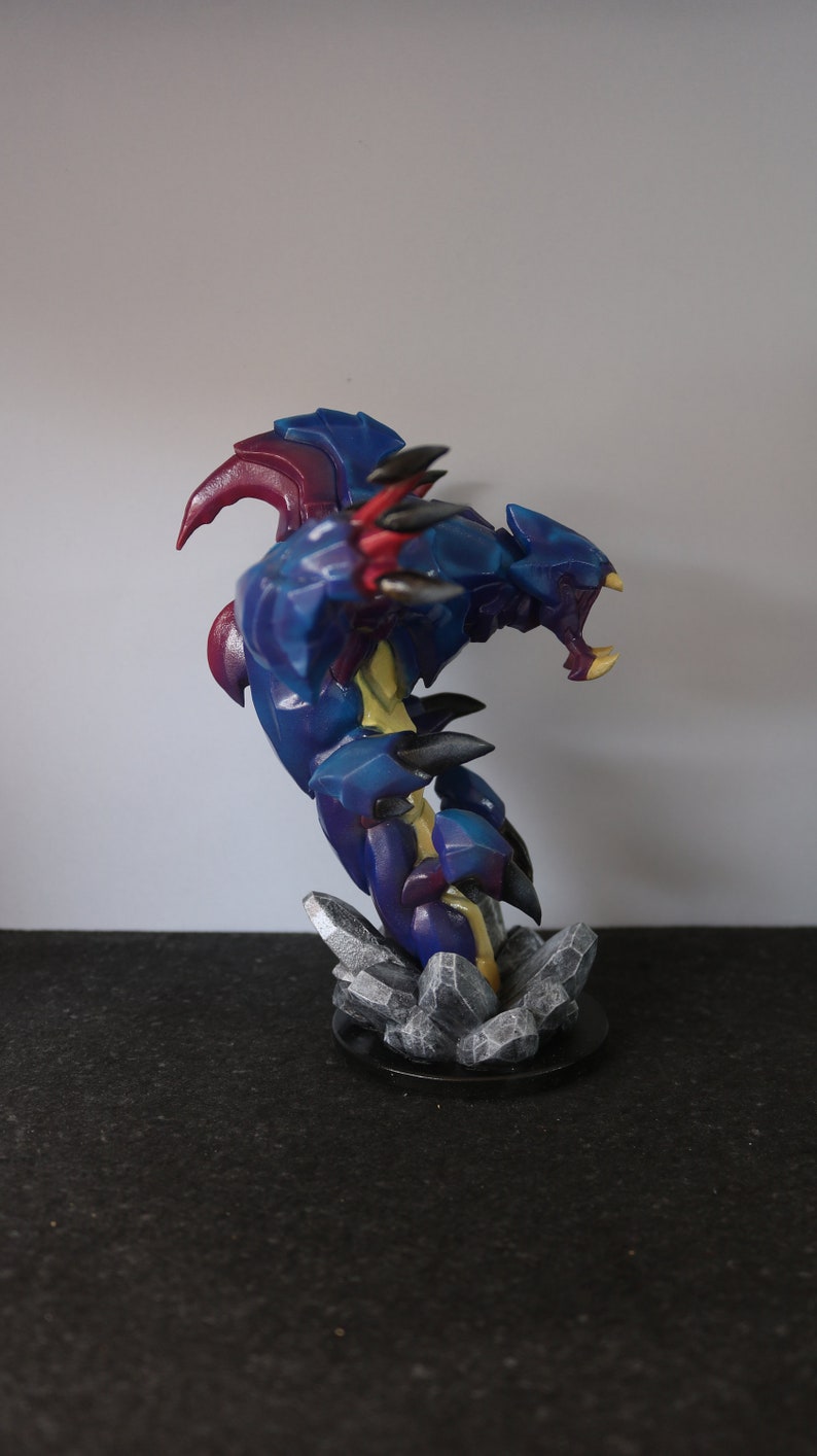 May include: A detailed figurine of a fantasy creature, predominantly blue with accents of red, yellow, and black. The sculpture is posed in an aggressive stance, mounted on a base of gray rock formations.