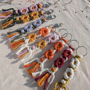 Könnte beinhalten: Ein Set aus bunten Makramee-Schlüsselanhängern mit Blumendesigns. Jeder Schlüsselanhänger hat eine andere Farbkombination und ein anderes Blumenmuster, darunter Orange, Gelb, Rosa, Weiß, Rot und Grün. Die Schlüsselanhänger sind aus verschiedenen farbigen Schnüren gefertigt und mit einem silbernen Schlüsselring versehen.