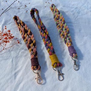 Op de afbeelding: Drie gevlochten sleutelhangers in verschillende kleuren. Elke sleutelhanger heeft een verschillend gekleurd uiteinde en een zilveren sluiting. De sleutelhangers liggen op een witte ondergrond.
