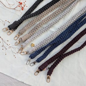 Könnte beinhalten: Eine Auswahl an Makramee-Taschenriemen in verschiedenen Farben wie Schwarz, Beige, Grau, Marineblau und Burgunderrot. Jeder Riemen hat ein gewebtes Design und goldfarbene Metallverschlüsse. Die Riemen liegen auf einer weißen Oberfläche.