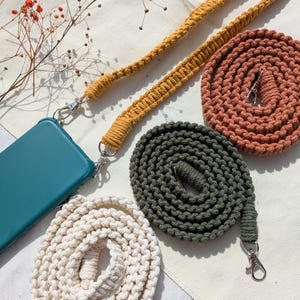 Könnte beinhalten: Vier Handy-Bänder in verschiedenen Farben: Creme, Senfgelb, Waldgrün und gebranntes Orange. Die Bänder sind aus gewebtem Baumwollkordel gefertigt und haben silberne Verschlüsse.