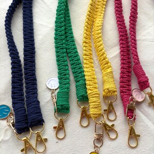 Könnte beinhalten: Vier Makramee-Lanyards in Marineblau, Grün, Gelb und Magenta. Jede Kordel hat eine geflochtene Textur und ist an goldfarbenen Karabinerverschlüssen aus Metall befestigt. Kleine runde Anhänger mit Smiley-Symbolen in passenden Farben hängen an den Verschlüssen.
