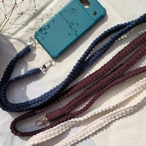 Könnte beinhalten: Türkisfarbene Handyhülle mit floralem Muster, kombiniert mit drei Makramee-Handy-Lanyards in Marineblau, Burgunderrot und Creme. Jedes Lanyard hat einen Metallverschluss. Die Handyhülle und Lanyards werden auf einem cremefarbenen Stoff präsentiert.