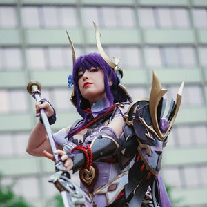 Puede incluir: Una mujer con un traje morado y negro con un casco dorado y morado sostiene una lanza larga de plata con una hoja azul. Lleva un atuendo morado y negro con detalles dorados. El traje es muy detallado y muy intrincado.