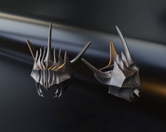 May include: Two silver and gold helmets with sharp points and intricate details. The helmets are on a dark background.