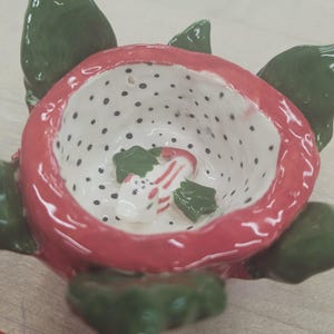 Pode incluir: Uma tigela de cerâmica em forma de morango, com exterior vermelho e interior branco salpicado de pontos pretos. A tigela contém uma pequena peça de cerâmica branca e vermelha e detalhes de folhas verdes.
