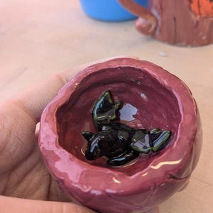 Pode incluir: Uma pequena tigela de cerâmica vermelha com um exterior áspero e texturizado. Dentro da tigela, há uma figura de morcego de cerâmica preta com asas verdes.