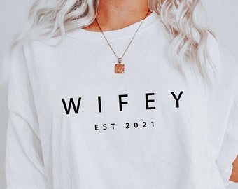 May include: White t-shirt with the word "WIFEY" printed in black, with "EST 2021" below it. The shirt is paired with light blue jeans. A gold necklace and bracelet are also visible.