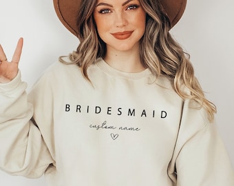 May include: A beige sweatshirt with the text "BRIDESMAID" and "custom name" in black lettering. The sweatshirt has a heart symbol below the text. A person is wearing the sweatshirt and a brown hat.