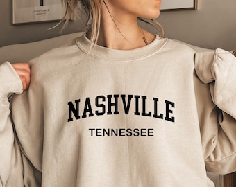 May include: A beige crewneck sweatshirt featuring the black text NASHVILLE TENNESSEE printed in a bold, arched serif font across the chest. The garment has long sleeves with ribbed cuffs and a ribbed neckline, providing a standard fit.
