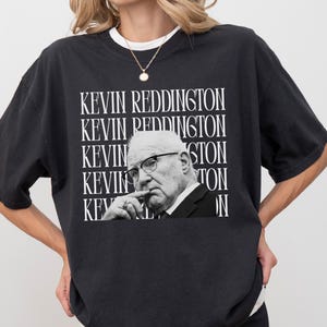 A woman wearing a black t-shirt with a graphic of a man's face and the text "KEVIN REDDINGTON" repeated across the shirt.