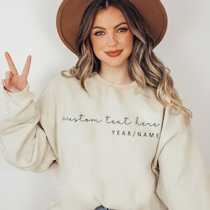 May include: A woman wearing a light beige sweatshirt with the text "custom text here YEAR/NAME" in black script. She is wearing a brown fedora and has long brown hair.