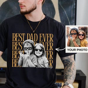 A man wearing a black t-shirt with a graphic design featuring two people and text, sitting on a couch.