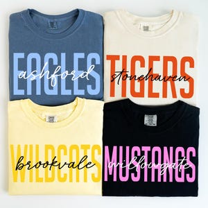 Puede incluir: Cuatro camisetas dobladas en azul, crema, amarillo y negro. Cada camiseta presenta un nombre de equipo en letras de bloque en negrita con un nombre en escritura m&aacute;s peque&ntilde;o debajo. Las camisetas est&aacute;n etiquetadas como "Eagles", "Tigers", "Wildcats" y "Mustangs".