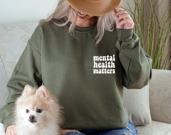 Puede incluir: Una mujer lleva una sudadera verde con el texto "mental health matters" en letras blancas. Lleva un sombrero marrón y unos vaqueros azules. Un pequeño perro blanco está sentado en su regazo.