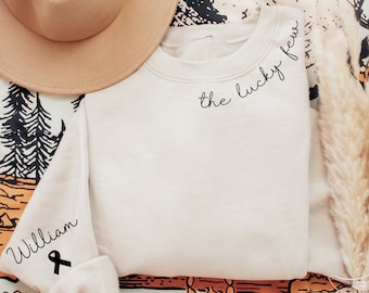 May include: A white sweatshirt with the text "the lucky few" on the collar and "William" with a black ribbon on the sleeve.