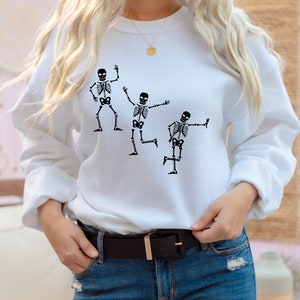 May include: White sweatshirt featuring a graphic of three skeletons dancing. The skeletons are black and the sweatshirt is white.