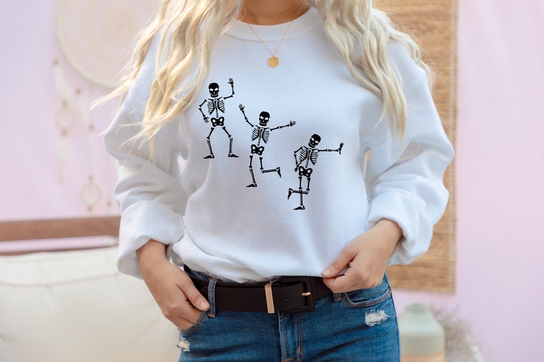 May include: White sweatshirt featuring a graphic of three skeletons dancing. The skeletons are black and the sweatshirt is white.