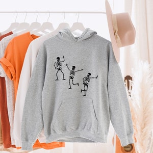 May include: A grey hoodie with a black print of three skeletons dancing. The hoodie has a drawstring hood and a kangaroo pocket.