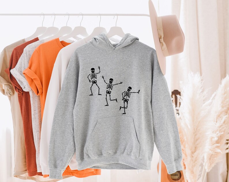 May include: A grey hoodie with a black print of three skeletons dancing. The hoodie has a drawstring hood and a kangaroo pocket.