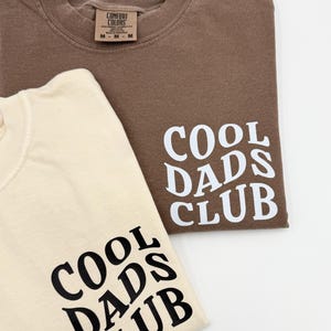 Pode incluir: Duas camisetas com o texto "Cool Dads Club" impresso nelas. Uma camiseta &eacute; marrom e a outra &eacute; bege.