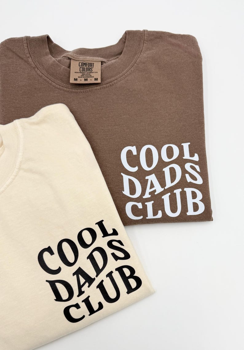 Pode incluir: Duas camisetas com o texto "Cool Dads Club" impresso nelas. Uma camiseta &eacute; marrom e a outra &eacute; bege.