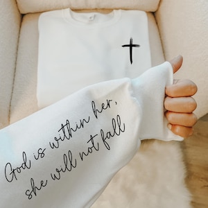 May include: White sweatshirt with a black cross on the front and the text "God is within her, she will not fall" on the sleeve.