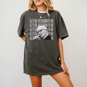 A woman wearing a gray t-shirt with a graphic of a man's face and the text "KEVIN REDDINGTON" repeated across the shirt.