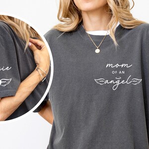 May include: Dark gray t-shirt with the words "mom of an angel" and a heart with wings graphic. The sleeve has the name "Jessie" and a similar heart and wing design. The shirt has a relaxed fit.
